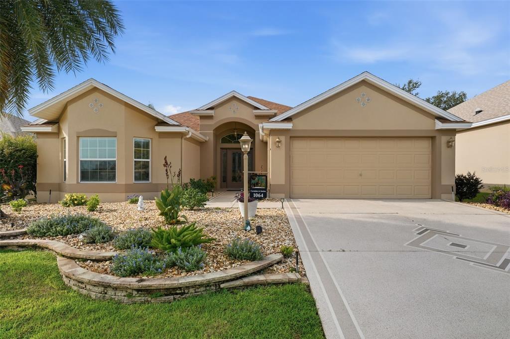 A single-story tan stucco house features a two-car garage and a landscaped front yard — 1064 ALCOVE LOOP, THE VILLAGES, FL