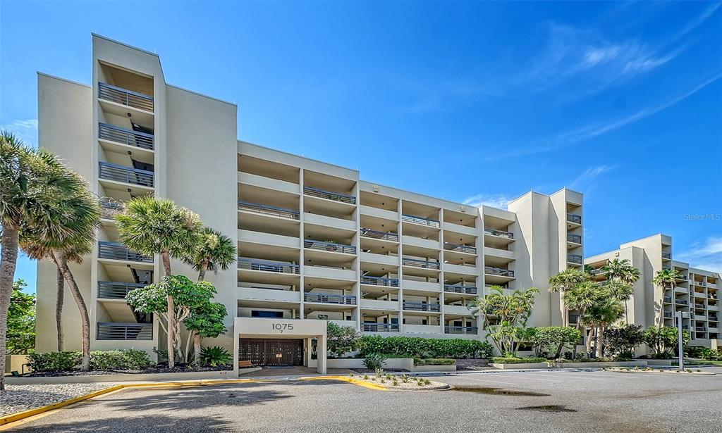 A multi-story apartment building with balconies and palm trees sits under a clear blue sky — 1075 GULF OF MEXICO DRIVE UNIT 303, LONGBOAT KEY, FL