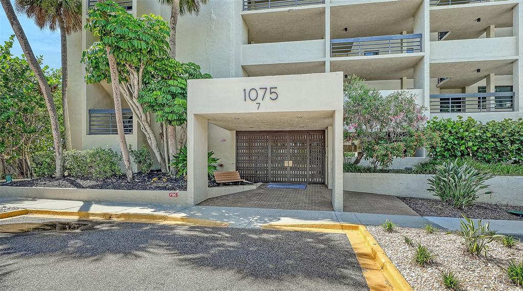The exterior entrance of a multi-story apartment building features a covered walkway and landscaping — 1075 GULF OF MEXICO DRIVE UNIT 303, LONGBOAT KEY, FL