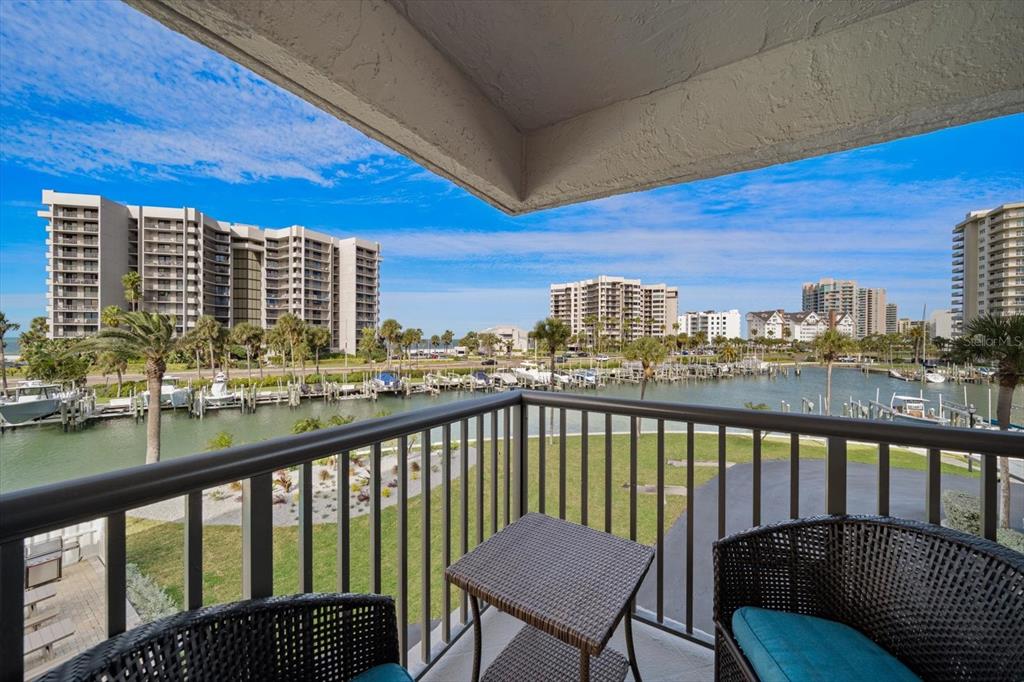 A balcony with wicker furniture overlooks a marina, palm trees, and several high-rise buildings — 1651 SAND KEY ESTATES COURT UNIT 28, CLEARWATER BEACH, FL