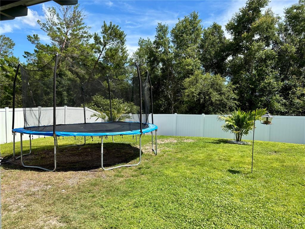 A backyard features a trampoline on the grass, a white vinyl fence, and mature trees — 3019 STATEN DRIVE, DELTONA, FL