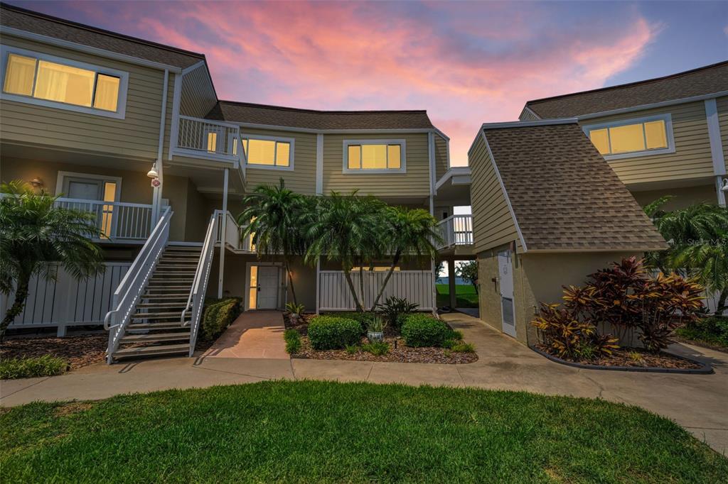 Exterior view of a multi-story tan apartment building with stairs and tropical landscaping at dusk — 433 S PAULA DRIVE UNIT 32, DUNEDIN, FL