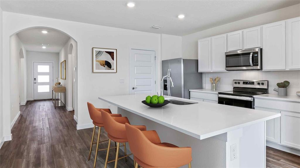 A modern kitchen features white cabinets, a large quartz island with orange stools, and stainless appliances — 11975 FALLOW DOWN DRIVE, DADE CITY, FL