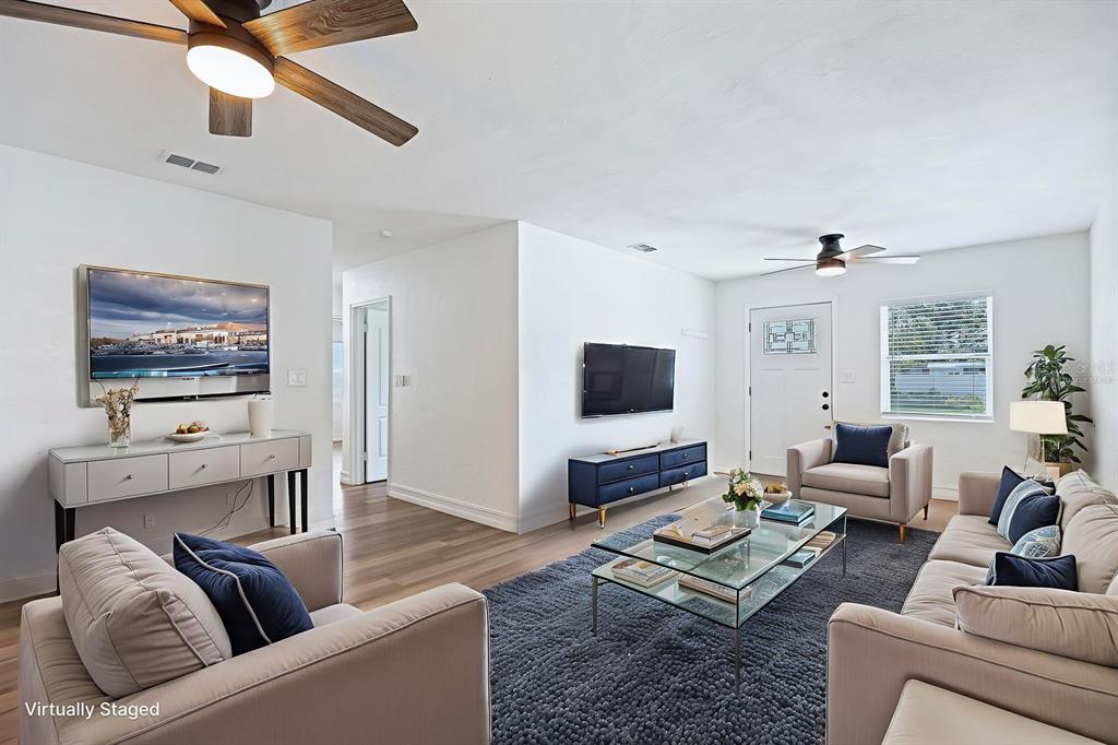 A living room with light wood floors, white walls, two ceiling fans, and virtual staging — 4006 S SHAMROCK ROAD, TAMPA, FL