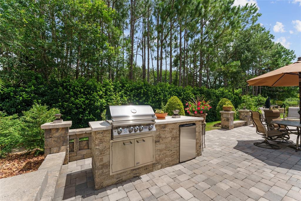 An outdoor kitchen with a stainless steel grill and refrigerator sits on a stone patio — 9158 SW 65TH LOOP, OCALA, FL