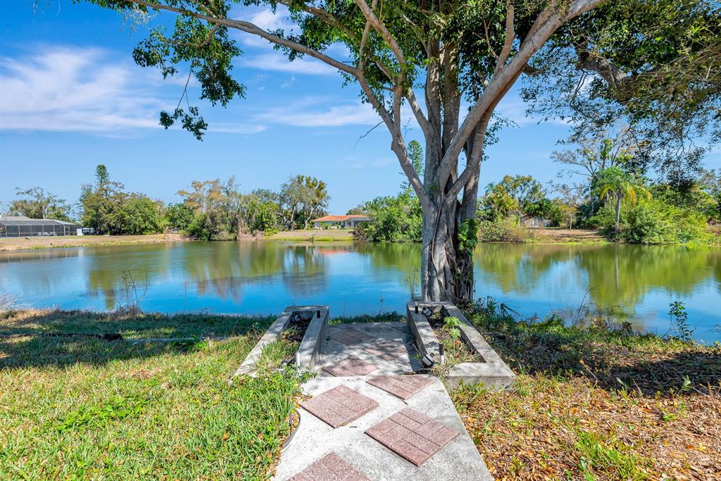 A concrete walkway leads toward a pond framed by a large tree and landscaping — 5017 80TH AVENUE CIRCLE E, SARASOTA, FL