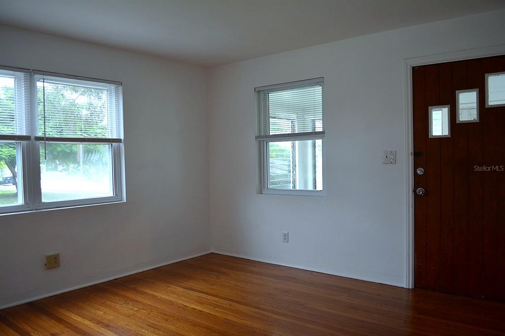 An empty room features hardwood floors, white walls, two windows, and a wooden front door — 1401 34TH AVENUE N, ST PETERSBURG, FL