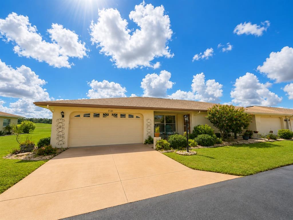 A single-story house with a beige garage, tan driveway, and green lawn under a cloudy sky — 310 FAIRCROSS CIRCLE, SUN CITY CENTER, FL