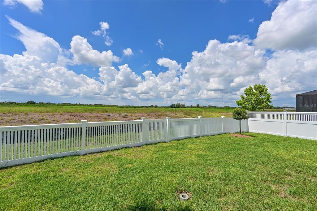 A white vinyl fence encloses a grassy backyard overlooking an open field under a cloudy sky — 3519 NIGHT STAR TERRACE, PARRISH, FL