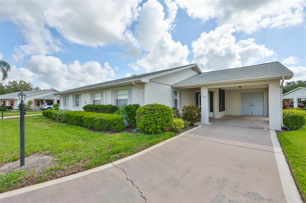 A one-story house with a concrete driveway, carport, and manicured landscaping under a cloudy sky — 109 GLENDOWER CIRCLE, SUN CITY CENTER, FL