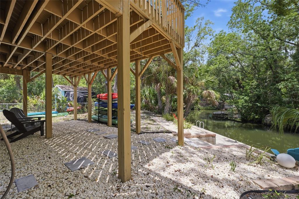 A gravel patio sits beneath a wooden deck overlooking a canal with stacked kayaks nearby — 8180 ANCHOR POINT DRIVE, WEEKI WACHEE, FL