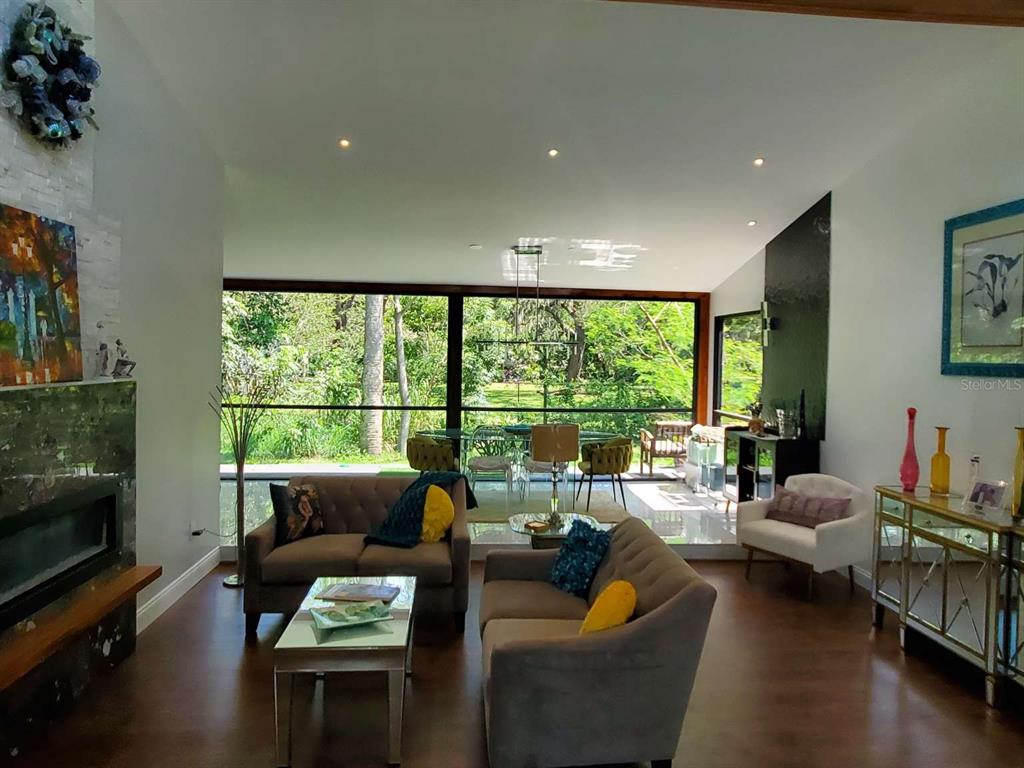 A living room features a fireplace, gray sofas, and a large window overlooking a garden — 5017 E 127TH AVENUE, TEMPLE TERRACE, FL