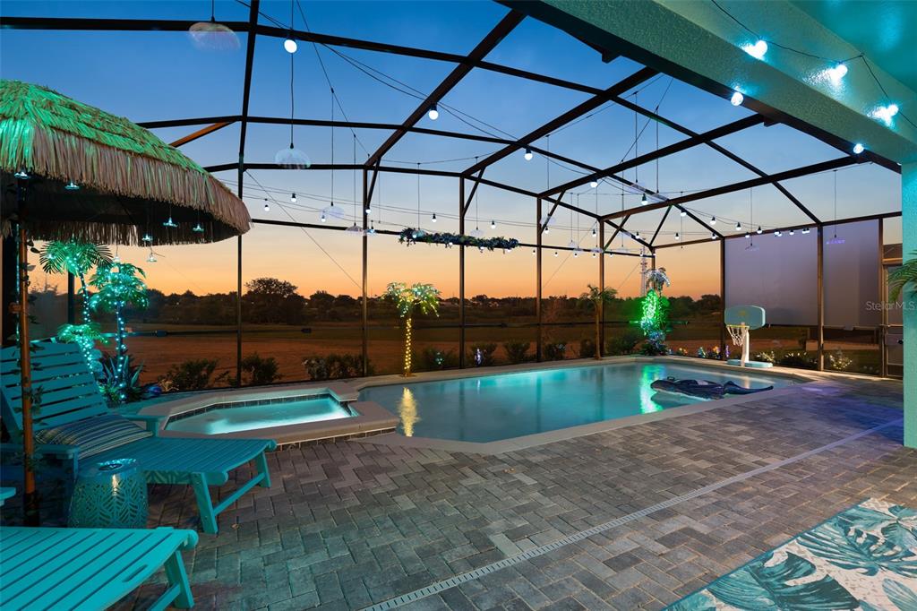 A screened-in pool patio features a hot tub, lounge chairs, and string lights at sunset — 16016 SANDY CLIFFS DRIVE, CLERMONT, FL