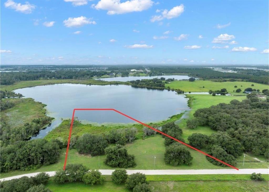 An aerial view shows a vacant grassy lot with red boundary lines bordering a lake — 9028 MCKENDREE ROAD, WESLEY CHAPEL, FL