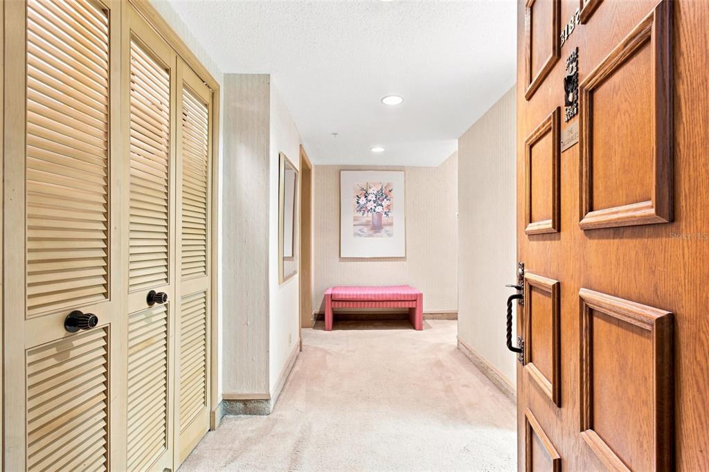 A hallway features beige carpet, louvered closet doors, a pink bench, and a wooden door — 36750 US HIGHWAY 19 UNIT 21105, PALM HARBOR, FL