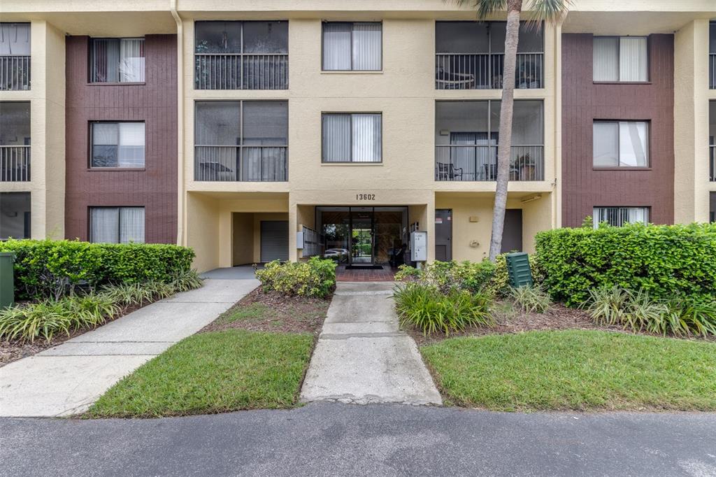 A three-story apartment building exterior with a central entrance, balconies, and surrounding landscaping — 13602 S VILLAGE DRIVE UNIT 1107, TAMPA, FL