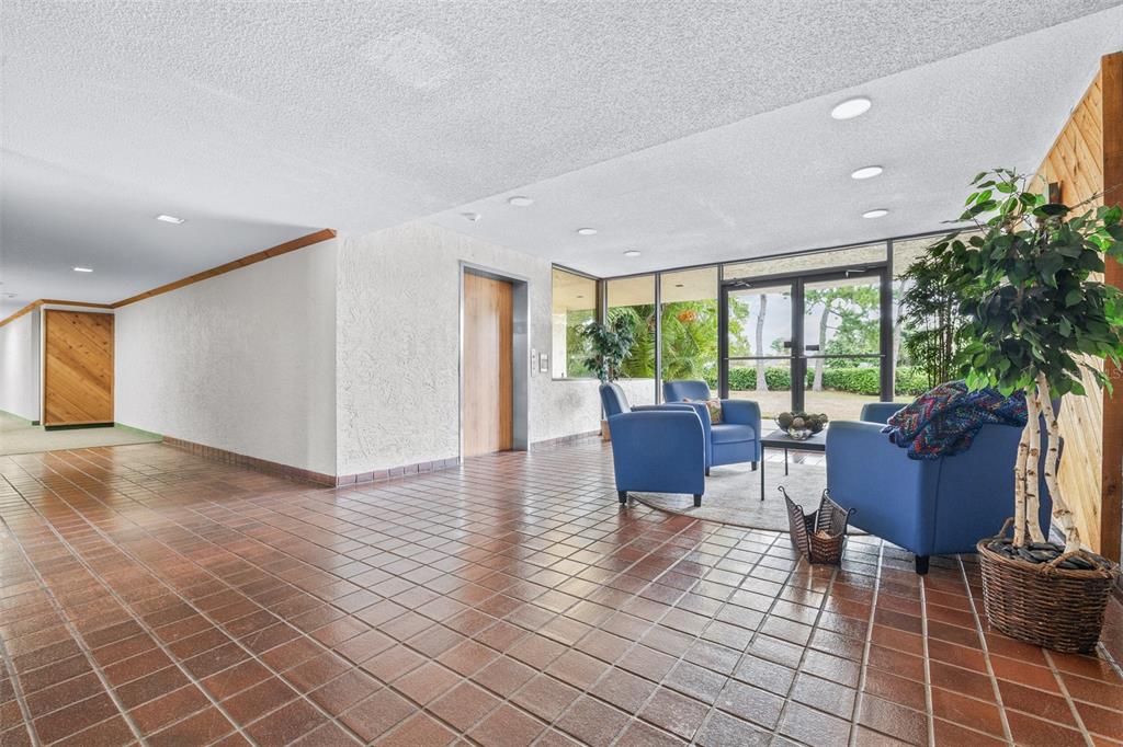 A lobby with brown tile floors, blue armchairs, a potted tree, and glass exterior doors — 13602 S VILLAGE DRIVE UNIT 1107, TAMPA, FL