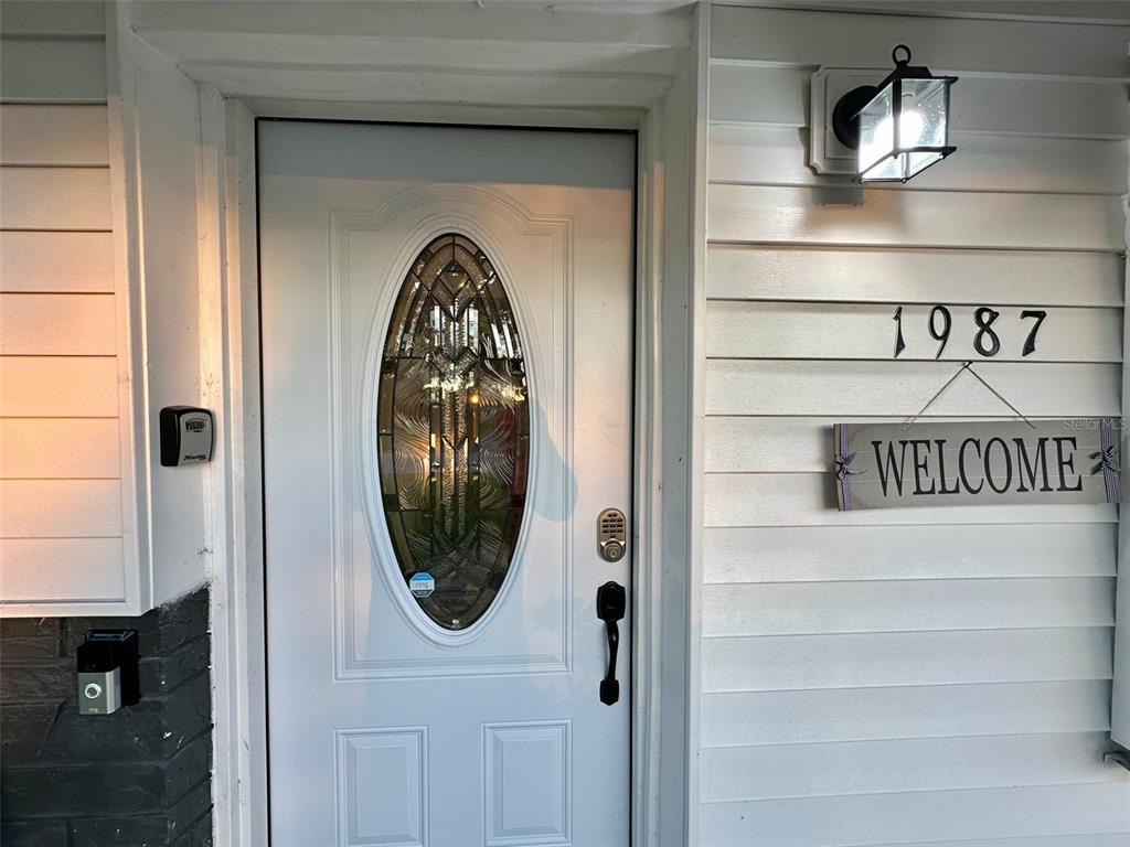 A white front door with an oval glass insert sits beside a welcome sign — 1987 MOSSWOOD DRIVE, MELBOURNE, FL