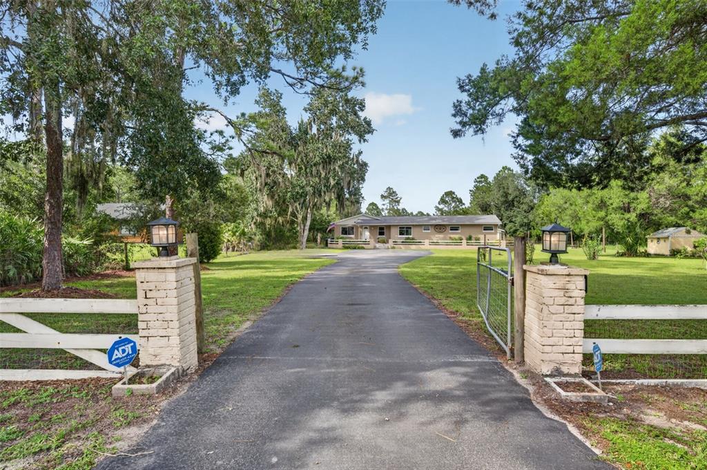 A paved driveway leads to a single-story house framed by trees and a gated entrance — 20963 E COLONIAL DRIVE, CHRISTMAS, FL