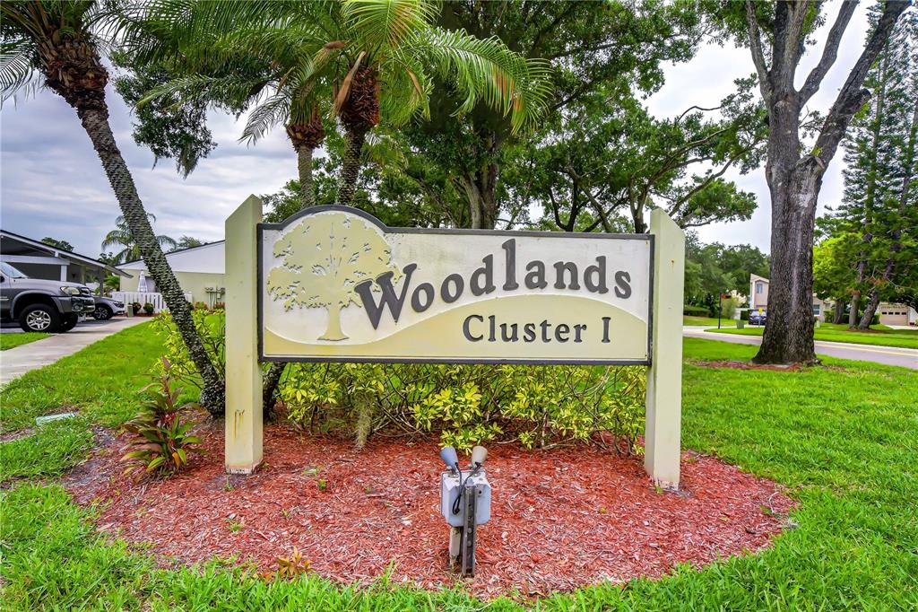 A sign for Woodlands Cluster I stands in a grassy area with trees — 230 CYPRESS PLACE, OLDSMAR, FL