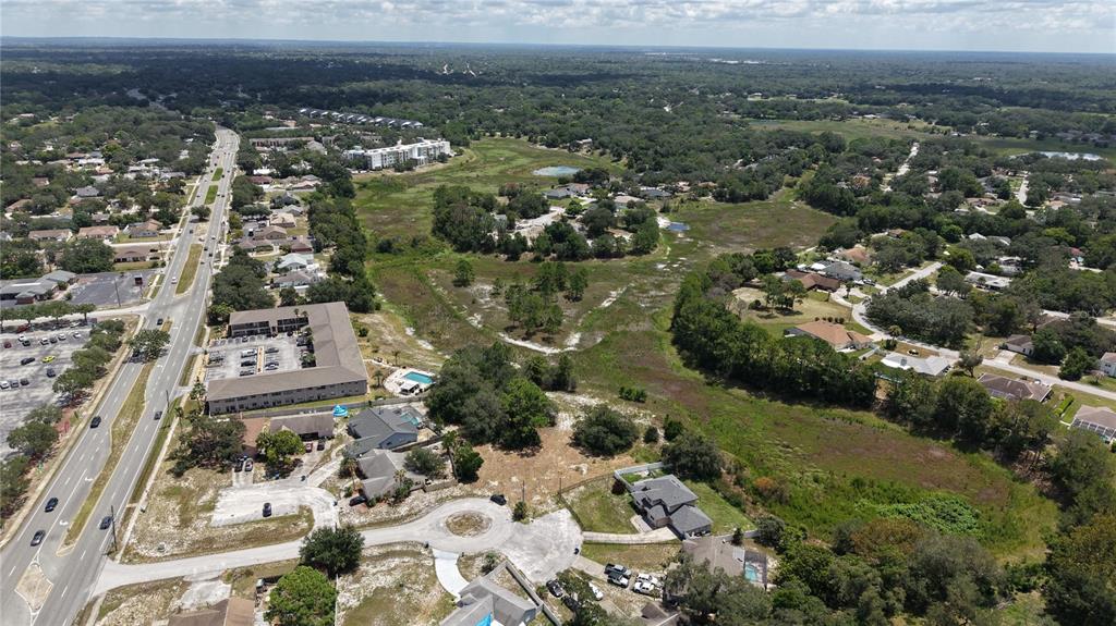 An aerial view shows a suburban neighborhood with houses, a multi-story building, and undeveloped land — 1170 PORT COURT, SPRING HILL, FL