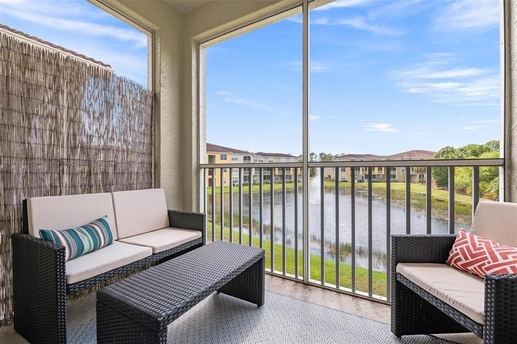 A screened-in balcony features wicker furniture overlooking a pond with a fountain and apartment buildings — 5681 BENTGRASS DRIVE UNIT 204, SARASOTA, FL