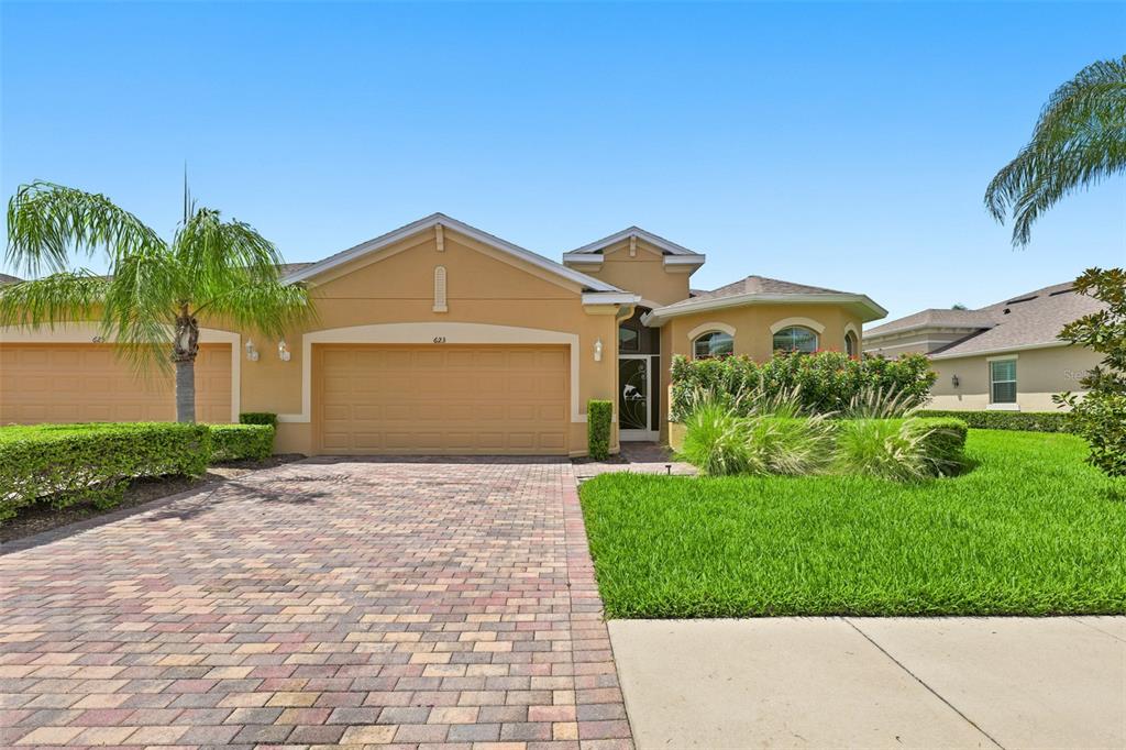 A single-story house with a tan stucco exterior, a two-car garage, and a brick driveway — 623 CHIPPER DRIVE, SUN CITY CENTER, FL
