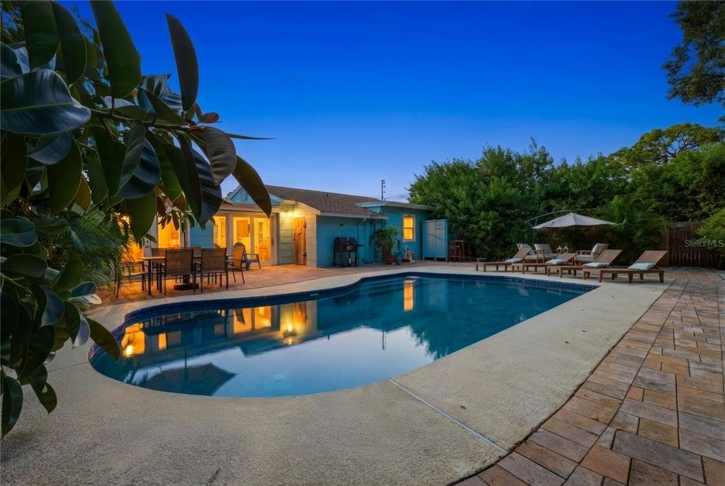 A backyard swimming pool with a patio, lounge chairs, and a blue house at dusk — 1249 ESSEX DRIVE N, ST PETERSBURG, FL