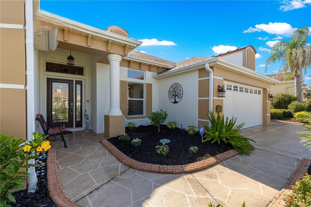 The exterior of a tan house features a covered entryway, a garage, and landscaped beds — 571 TROY LOOP, THE VILLAGES, FL