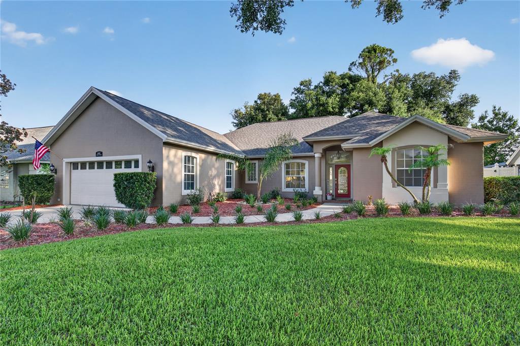 A single-story house with a beige stucco exterior, a two-car garage, and a red front door — 19337 SPRING OAK DRIVE, EUSTIS, FL