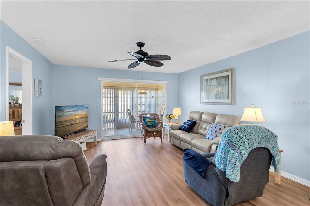 A living room features light blue walls, wood-look flooring, a ceiling fan, and sliding glass doors — 1701 PINEHURST ROAD UNIT 6A, DUNEDIN, FL