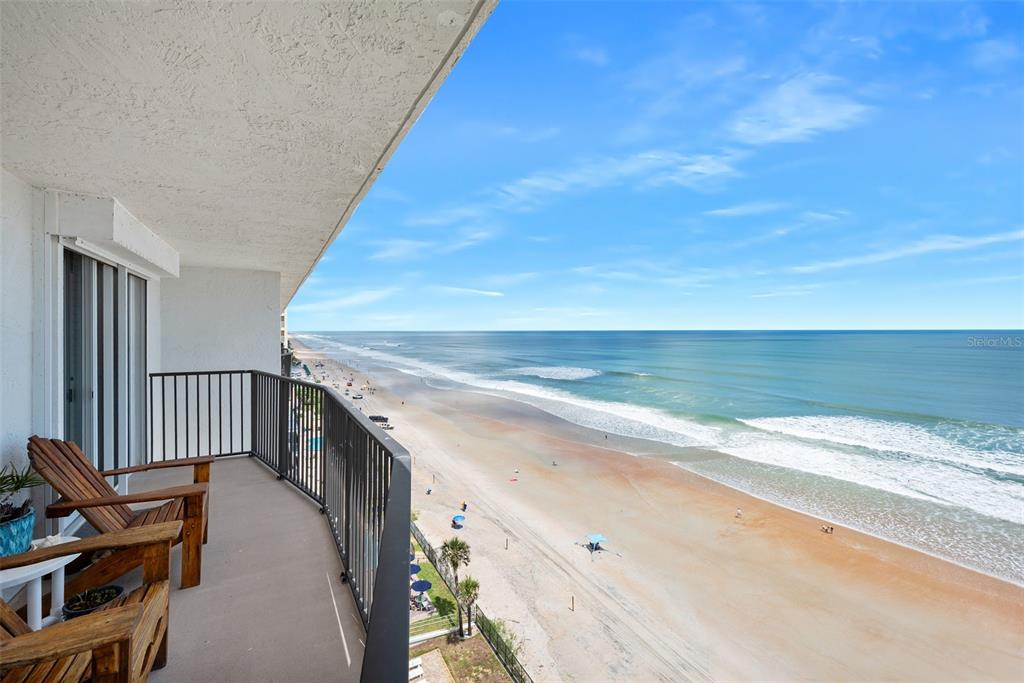A balcony with wooden chairs overlooks a sandy beach and the ocean under a blue sky — 111 S ATLANTIC AVENUE UNIT 1104, ORMOND BEACH, FL