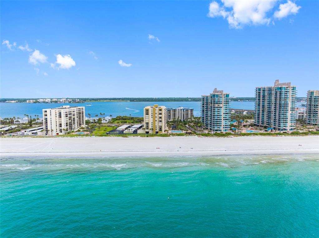 An aerial view shows high-rise buildings along a white sand beach and turquoise ocean — 1480 GULF BOULEVARD UNIT 1011, CLEARWATER BEACH, FL