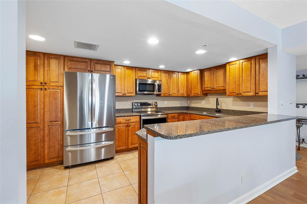 A kitchen features wood cabinets, stainless steel appliances, granite countertops, and a breakfast bar — 1480 GULF BOULEVARD UNIT 1011, CLEARWATER BEACH, FL