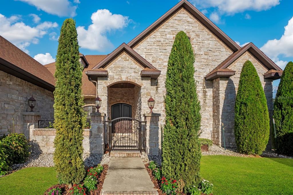 A stone-clad house exterior features a gated courtyard entrance flanked by tall, slender evergreen trees — 2310 NEVADA ROAD, LAKELAND, FL
