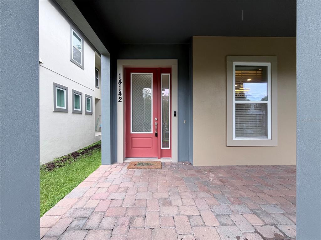 A red front door with a glass panel sits under a covered porch with pavers — 14142 WALCOTT AVENUE, ORLANDO, FL