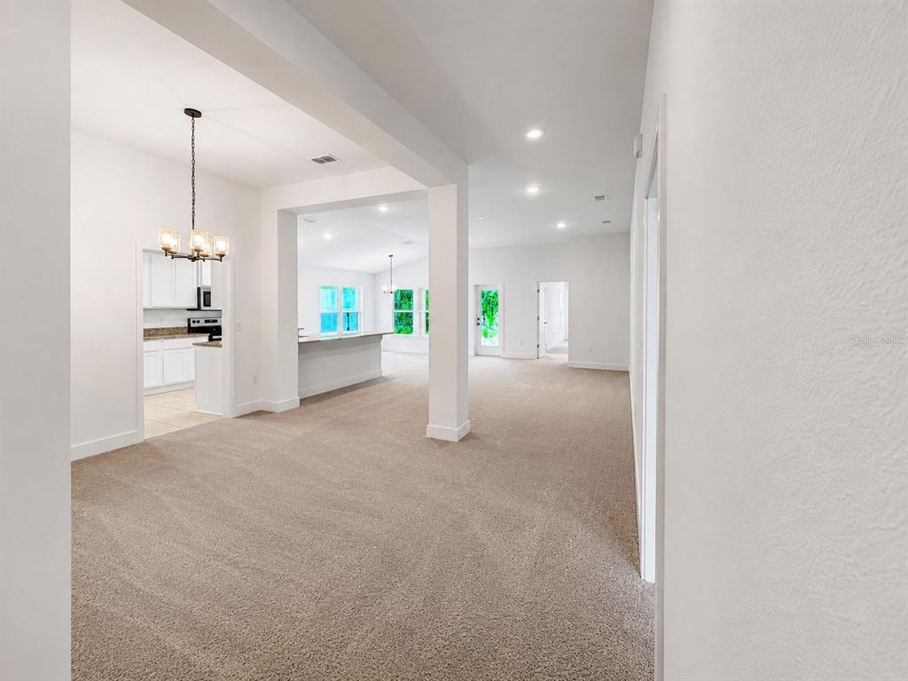 An open-concept living area with beige carpeting, white walls, and a view into the kitchen — 7618 BENT TREE LOOP, HARMONY, FL