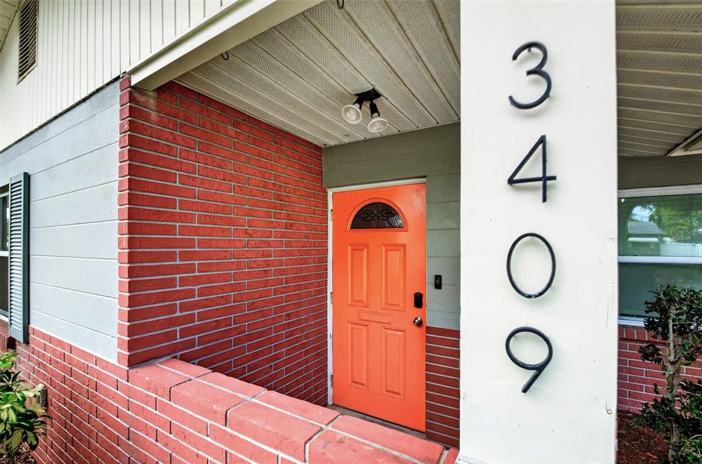 A bright orange front door is set against a red brick wall under a porch — 3409 18TH AVENUE DRIVE W, BRADENTON, FL