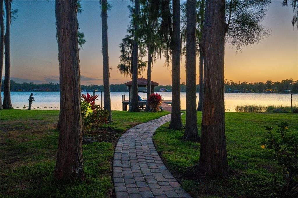 A brick path leads through trees toward a gazebo on a lake at sunset — 18258 WAYNE ROAD, ODESSA, FL