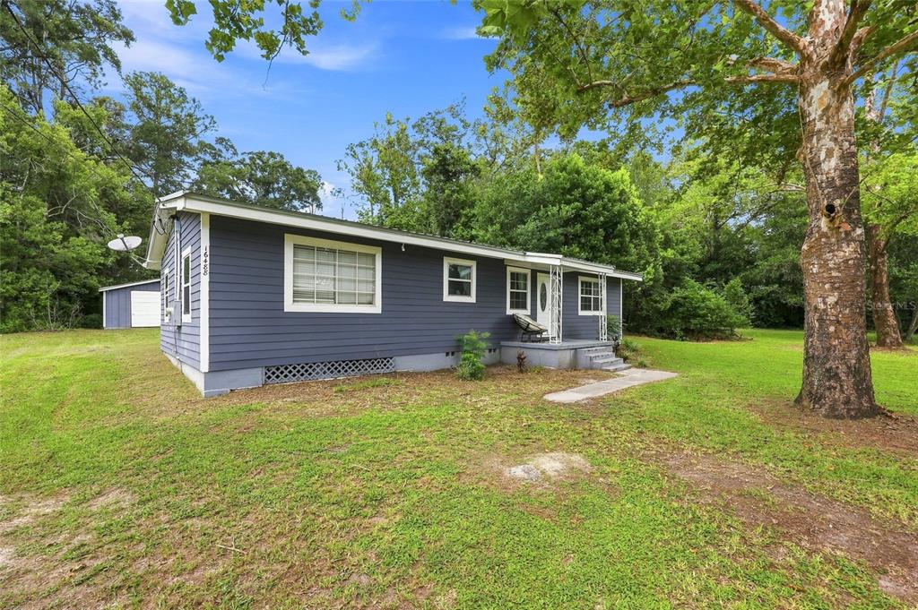 A blue, single-story house with a metal roof sits on a grassy, tree-filled lot — 16488 SW 139TH AVENUE, ARCHER, FL
