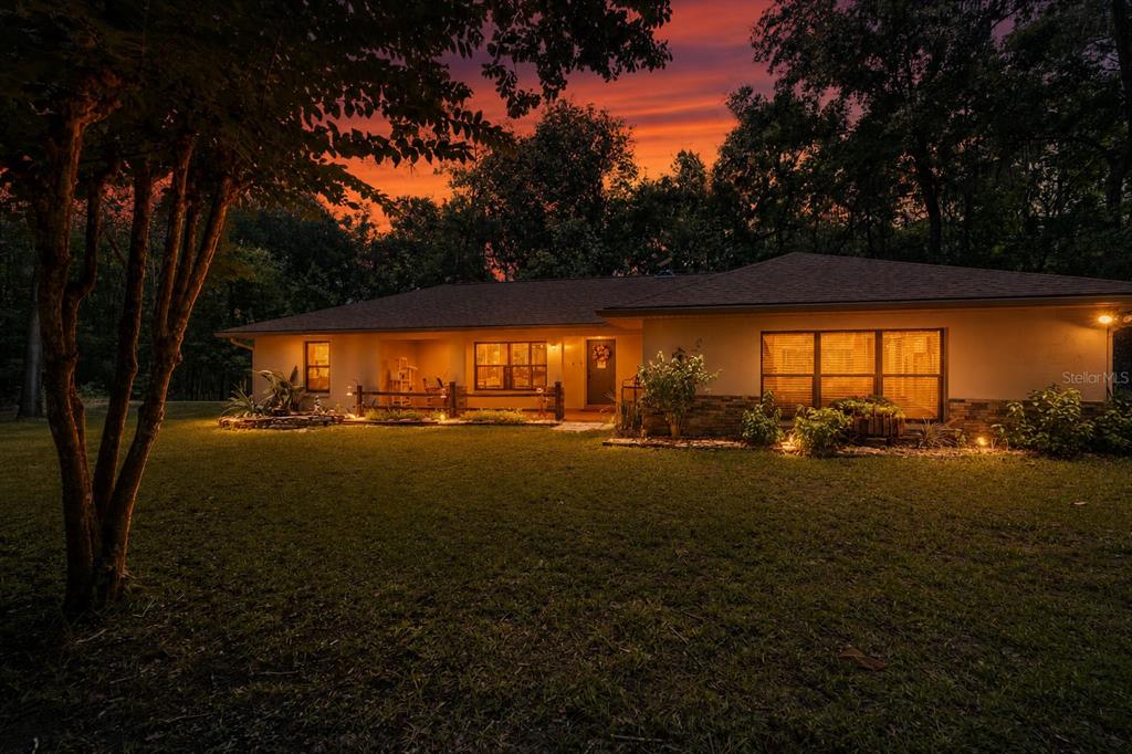 A single-story house with a stone-accented facade is illuminated at dusk under a sunset sky — 18451 NW 141ST AVENUE, WILLISTON, FL
