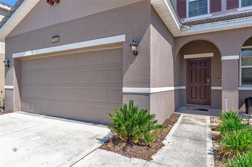A tan stucco house exterior features a two-car garage, a brown front door, and landscaping — 7635 OAKMOSS LOOP, DAVENPORT, FL