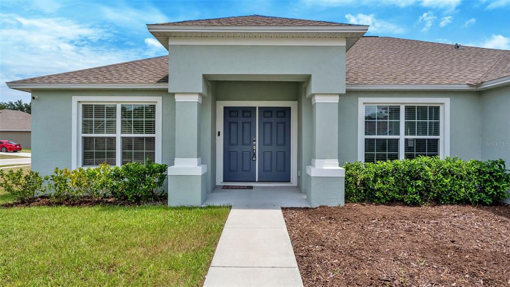 The exterior of a single-story house features a light green facade, dark blue double doors, and landscaping — 2048 WINDING RIVER COVE, BARTOW, FL