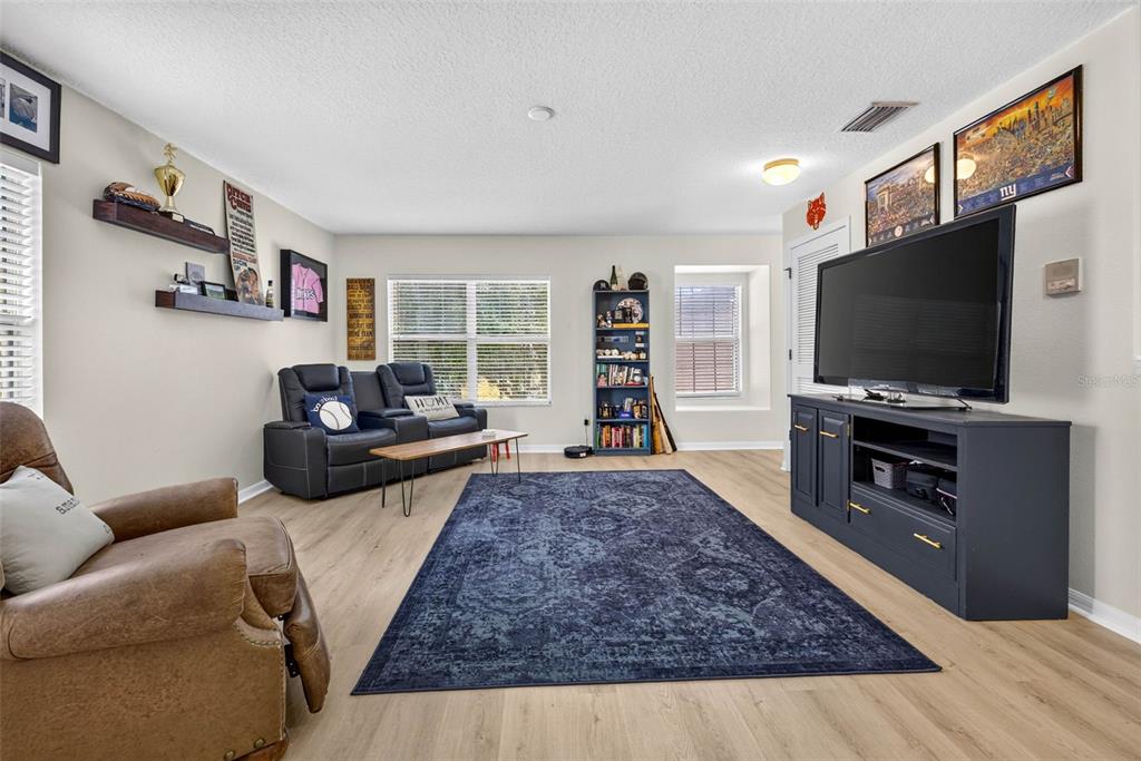 A living room features light wood flooring, a large blue area rug, and a television console — 7360 BRIDGEVIEW, WESLEY CHAPEL, FL