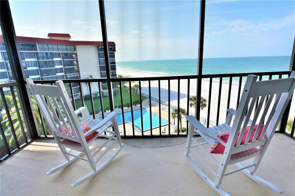 A screened balcony features two white rocking chairs overlooking a swimming pool and the ocean — 18304 GULF BOULEVARD UNIT 504, REDINGTON SHORES, FL