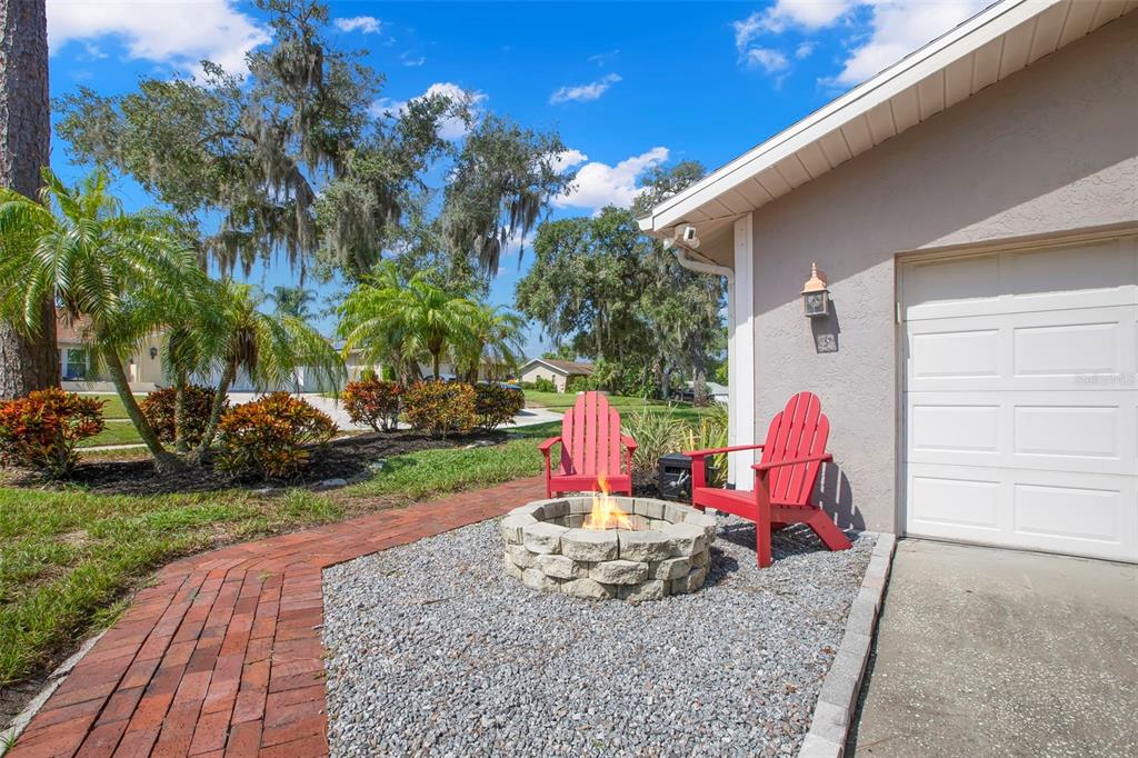 A stone fire pit with two red chairs sits on a gravel patio beside a garage — 1419 INDIAN TRAIL S, PALM HARBOR, FL