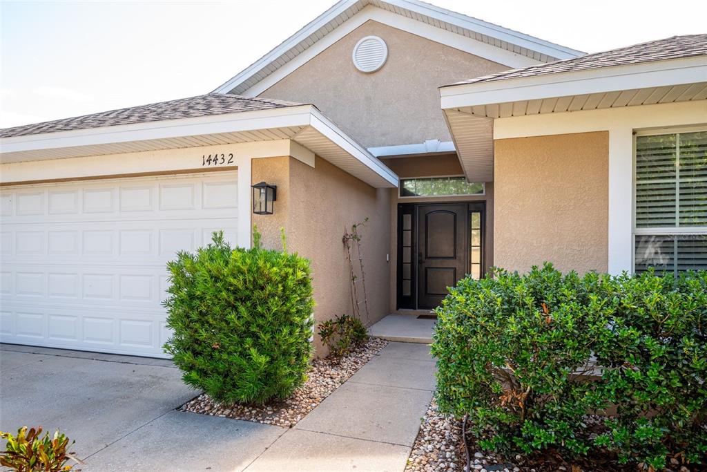 A tan house exterior features a white garage door, a front entrance, and green landscaping — 14432 BARLEY FIELD DRIVE, WIMAUMA, FL