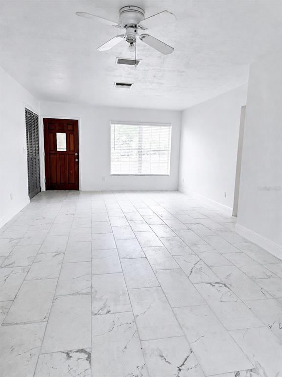 A bright, empty room features white marble-look tile flooring, a ceiling fan, and a wooden door — 1315 E HANCOCK DRIVE, DELTONA, FL