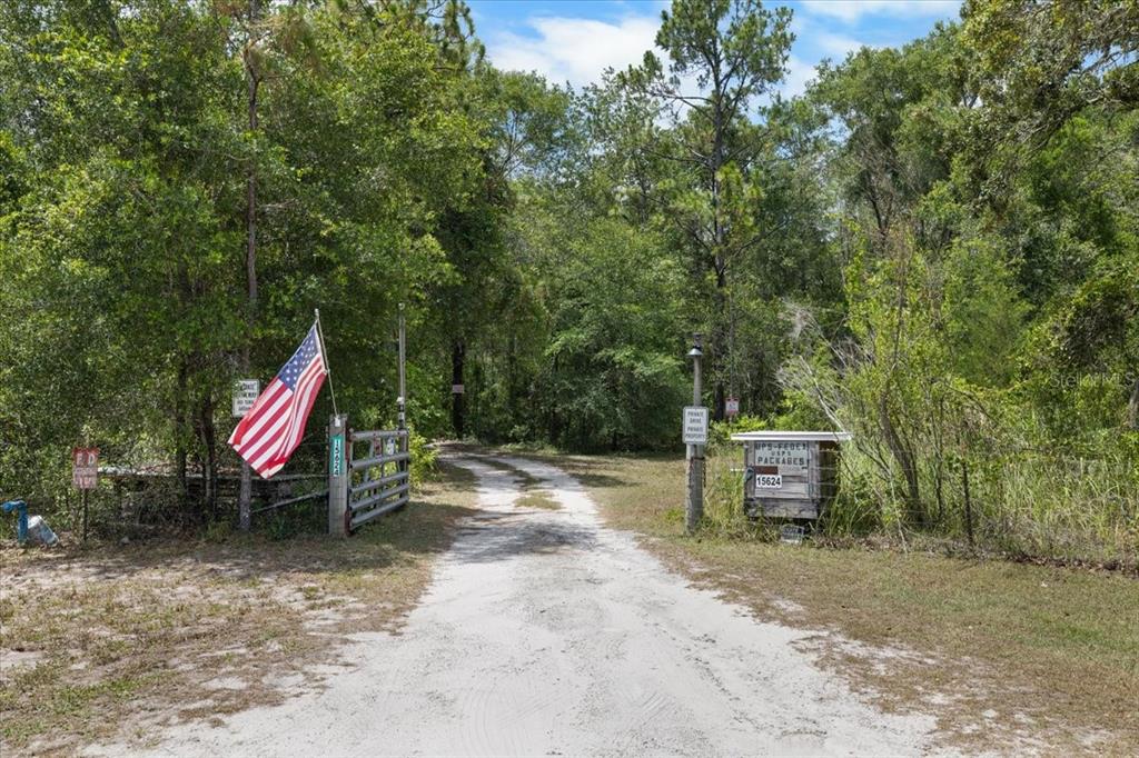 A dirt driveway leads through a gated entrance into a wooded area with an American flag — 15624 PEACE BOULEVARD, SPRING HILL, FL