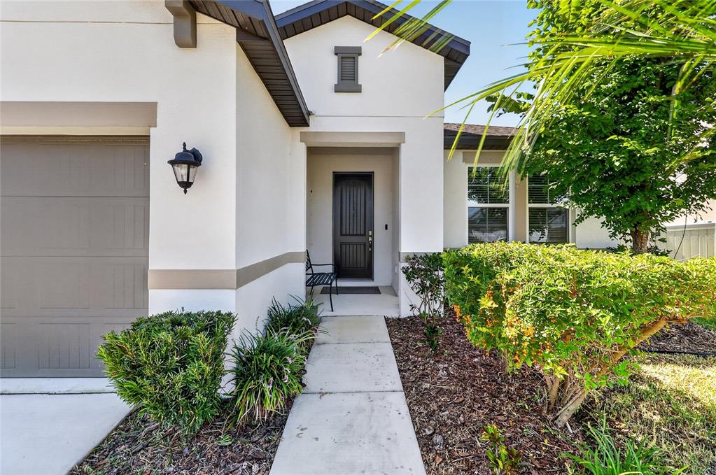A white stucco house exterior features a gray garage door, front entryway, and landscaped garden — 13520 PALMERA VISTA DRIVE, RIVERVIEW, FL